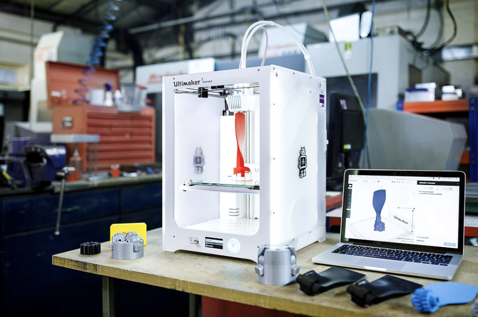 Domowa drukarka 3D (model Ultimaker 3 Extended) podczas drukowania prototypu z filamentem PLA; obok drukarki widać wydrukowane elementy oraz laptop z projektem modelu 3D